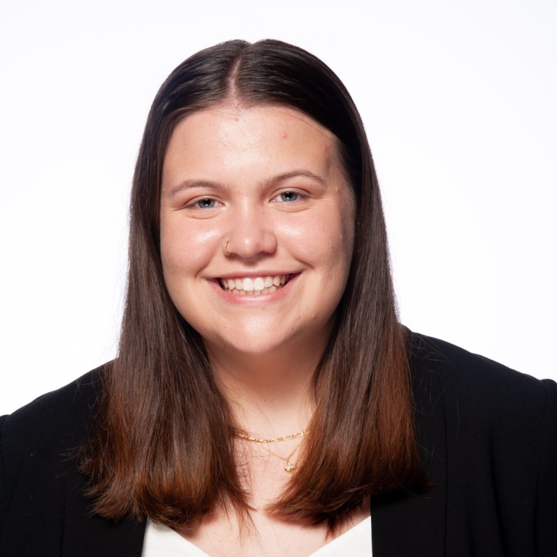 White woman with shoulder length brown hair, wearing a white shirt with a black blazer (Website owner)